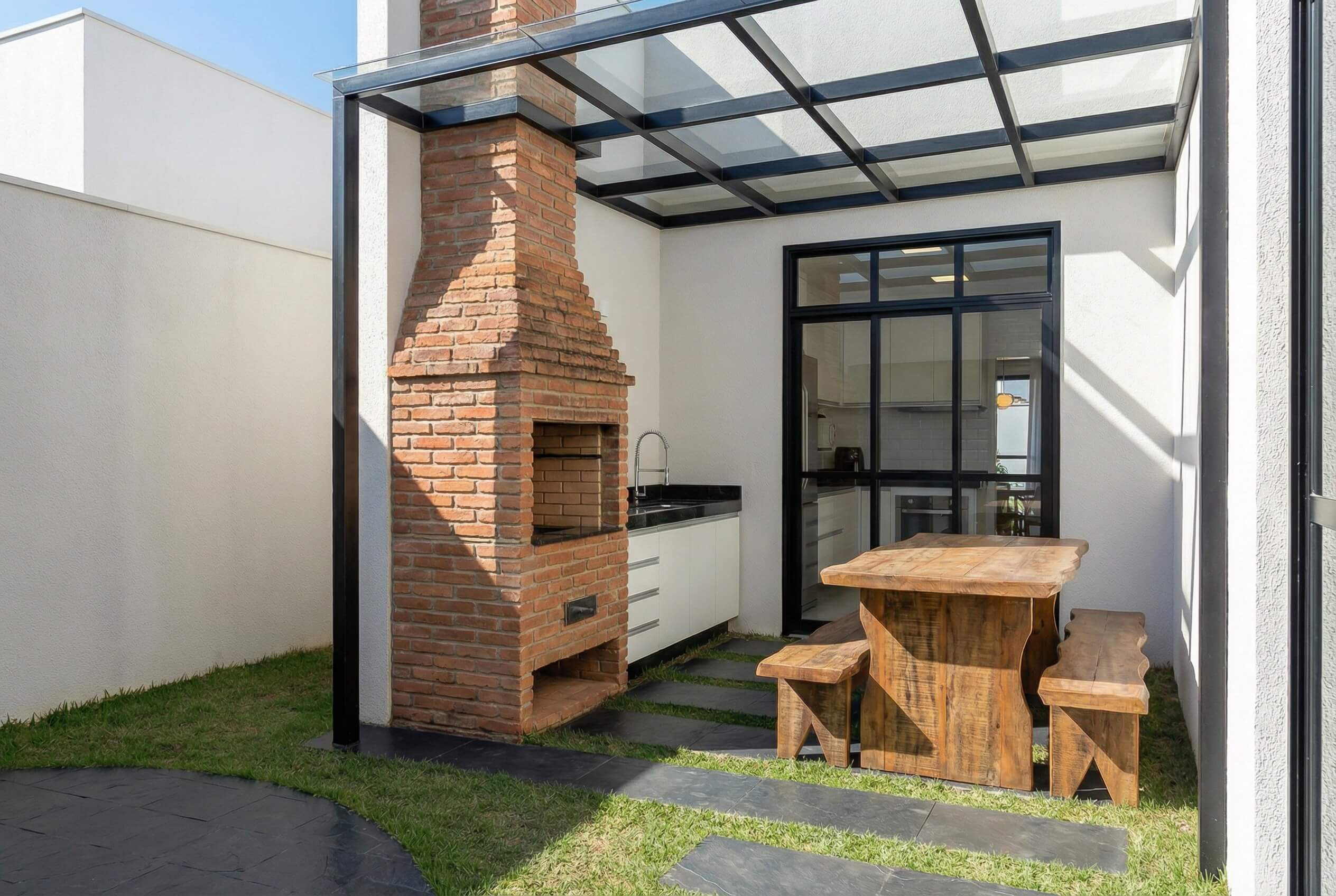 Área gourmet externa com churrasqueira de tijolos, cobertura de vidro e mesa de madeira